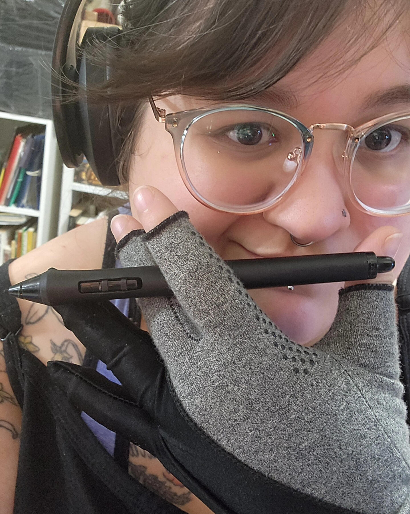 A light skinned Latine with brown eyes, large round glasses, a star nostril piercing, a silver septum ring, and lower lip stud. They're holding their hand up in front of their face, holding a digital art pen, while wearing both compression glove and drawin
